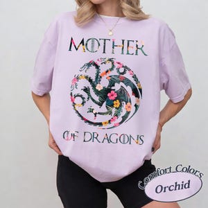 May include: A light purple short-sleeve t-shirt featuring a circular three-headed dragon graphic filled with a tropical floral pattern. The text MOTHER OF DRAGONS is printed in a dark font above and below the dragon emblem on the front.