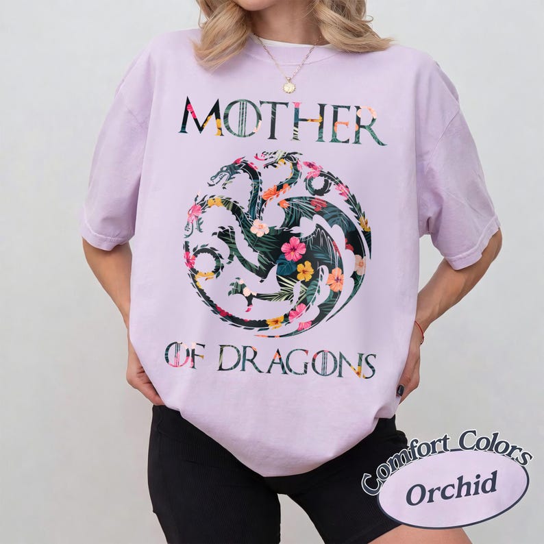 May include: A light purple short-sleeve t-shirt featuring a circular three-headed dragon graphic filled with a tropical floral pattern. The text MOTHER OF DRAGONS is printed in a dark font above and below the dragon emblem on the front.