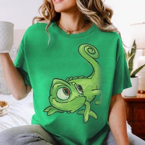 Peut inclure: Un t-shirt vert vif à manches courtes orné d'une grande illustration graphique détaillée d'un caméléon vert avec une queue enroulée et de grands yeux au centre. Le t-shirt présente une encolure ronde classique et une coupe décontractée.