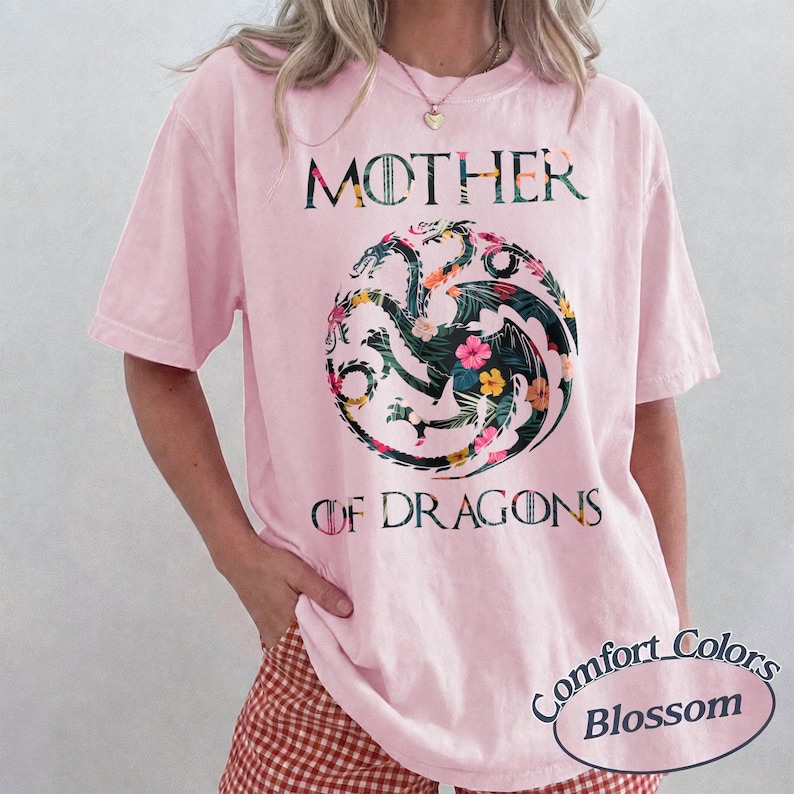 May include: A light pink short-sleeve t-shirt featuring a circular three-headed dragon graphic filled with colourful tropical flowers and leaves. The text MOTHER OF DRAGONS is printed in a dark font above and below the dragon illustration.