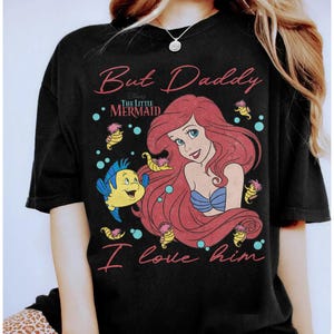 May include: A black t-shirt featuring a graphic of Ariel from The Little Mermaid with red hair, a blue seashell top, and Flounder the fish. The text reads But Daddy I love him in pink script, with small yellow seahorses and blue bubbles.