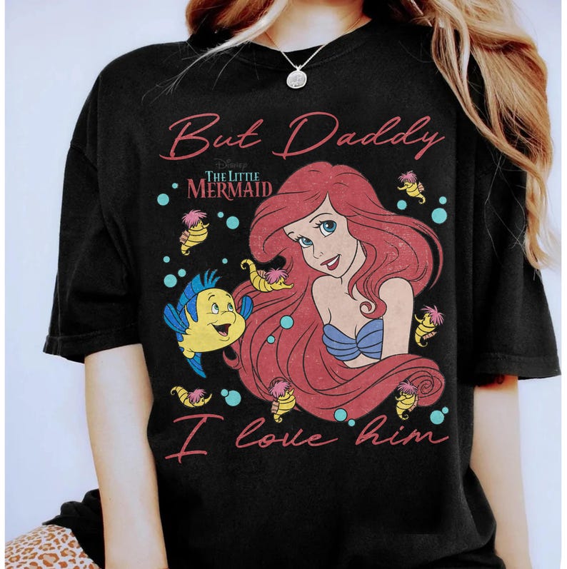 May include: A black t-shirt featuring a graphic of Ariel from The Little Mermaid with red hair, a blue seashell top, and Flounder the fish. The text reads But Daddy I love him in pink script, with small yellow seahorses and blue bubbles.