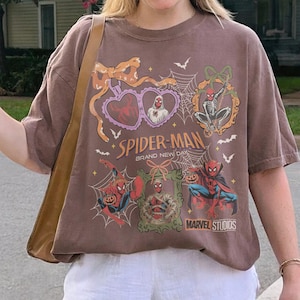 May include: A brown short-sleeve t-shirt featuring a graphic print with the text Spider-Man Brand New Day and Marvel Studios. The design includes illustrations of Spider-Man characters in Halloween-themed frames, spiderwebs, bats, and bows.