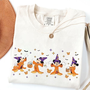 以下が含まれることがあります： ハロウィーンをテーマにした、ミッキーマウスのキャラクターが魔法使いに扮したグラフィックが特徴のクリーム色のTシャツ。オレンジ色のローブ、紫色の帽子、カボチャ型のスイーツのデザイン。金のネックレスと茶色の帽子も見える。