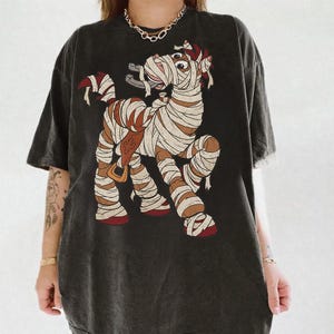 May include: A charcoal gray short-sleeve t-shirt featuring a graphic print of a brown horse wrapped in white bandages like a mummy. The horse is wearing a brown saddle and has a horseshoe in its mouth. The shirt has a crew neckline and a loose fit.
