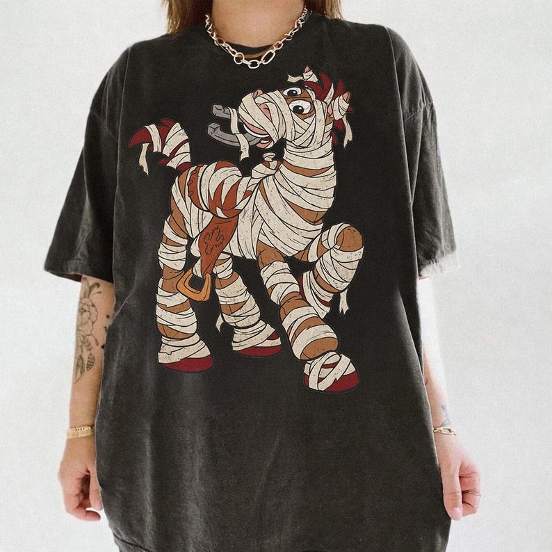 May include: A charcoal gray short-sleeve t-shirt featuring a graphic print of a brown horse wrapped in white bandages like a mummy. The horse is wearing a brown saddle and has a horseshoe in its mouth. The shirt has a crew neckline and a loose fit.