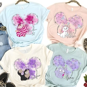 Puede incluir: Cuatro camisetas de manga corta en azul claro, rosa, crema y verde menta, cada una con un diseño de gato de dibujos animados dentro de un contorno de oreja de Mickey Mouse. Las camisetas tienen diferentes personajes de gatos y nombres impresos. Las camisetas se muestran sobre una superficie blanca.
