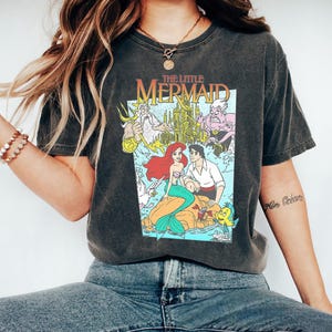 Puede incluir: Camiseta gris oscuro con un gráfico colorido de "La Sirenita". El diseño incluye a Ariel, el Príncipe Eric y otros personajes de la película. La camiseta tiene un aspecto vintage y desgastado.