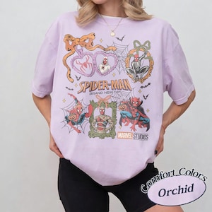 May include: A light purple t-shirt featuring a graphic print of Spider-Man in various Halloween-themed costumes and poses. The design includes spider webs, bats, bows, and frames, with the text Spider-Man Brand New Day and Marvel Studios.