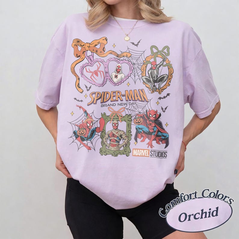 May include: A light purple t-shirt featuring a graphic print of Spider-Man in various Halloween-themed costumes and poses. The design includes spider webs, bats, bows, and frames, with the text Spider-Man Brand New Day and Marvel Studios.