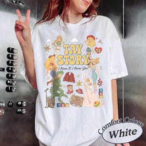 May include: A white t-shirt featuring a colourful graphic print with the text "TAY STORY" and "I Knew It, I Knew You". The design includes multiple illustrated figures, a red cardigan, cowboy boots, a hat, blocks, and a sign reading "TAY'S ROOM KEEP OUT".