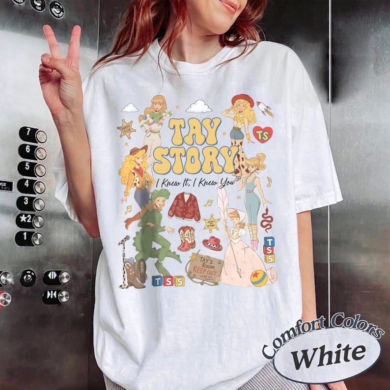 May include: A white t-shirt featuring a colourful graphic print with the text "TAY STORY" and "I Knew It, I Knew You". The design includes multiple illustrated figures, a red cardigan, cowboy boots, a hat, blocks, and a sign reading "TAY'S ROOM KEEP OUT".