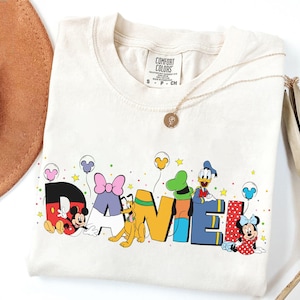 May include: Cream-colored t-shirt featuring the name "DANIEL" in colorful block letters with Disney characters. Mickey Mouse, Minnie Mouse, Donald Duck, and Pluto are depicted. The shirt has a round neckline and short sleeves.