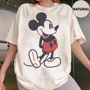 Könnte beinhalten: Cremefarbenes T-Shirt mit großem Mickey Mouse-Motiv. Die Zeichentrickfigur ist in Schwarz, Rot und Weiß dargestellt und lächelt. Das T-Shirt hat kurze Ärmel und eine lockere Passform. Ein kreisförmiges "NATURAL"-Etikett ist sichtbar.