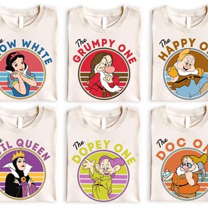 Puede incluir: Seis camisetas de color crema con gráficos circulares de Blancanieves, Gruñón, Feliz, la Reina Malvada, Mudito y Sabio. Cada camiseta muestra el nombre del personaje en una fuente retro sobre un fondo de atardecer colorido en un marco circular.
