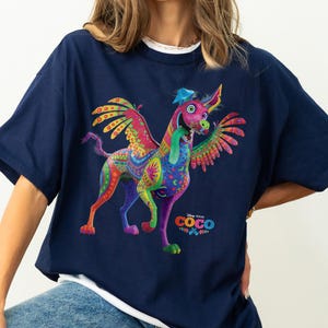 May include: A navy blue short-sleeve t-shirt featuring a colorful graphic of Dante the alebrije dog from the movie Coco. The dog has wings, a long green tongue, and intricate patterns in shades of pink, yellow, and green. The Disney Pixar Coco logo is printed below the dog.