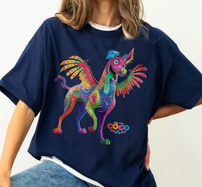May include: A navy blue short-sleeve t-shirt featuring a colorful graphic of Dante the alebrije dog from the movie Coco. The dog has wings, a long green tongue, and intricate patterns in shades of pink, yellow, and green. The Disney Pixar Coco logo is printed below the dog.