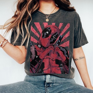 May include: A charcoal gray short-sleeve t-shirt featuring a graphic print of the character Deadpool making a heart shape with their hands. The design includes a red sunburst background with black halftone dots and two sword hilts visible.