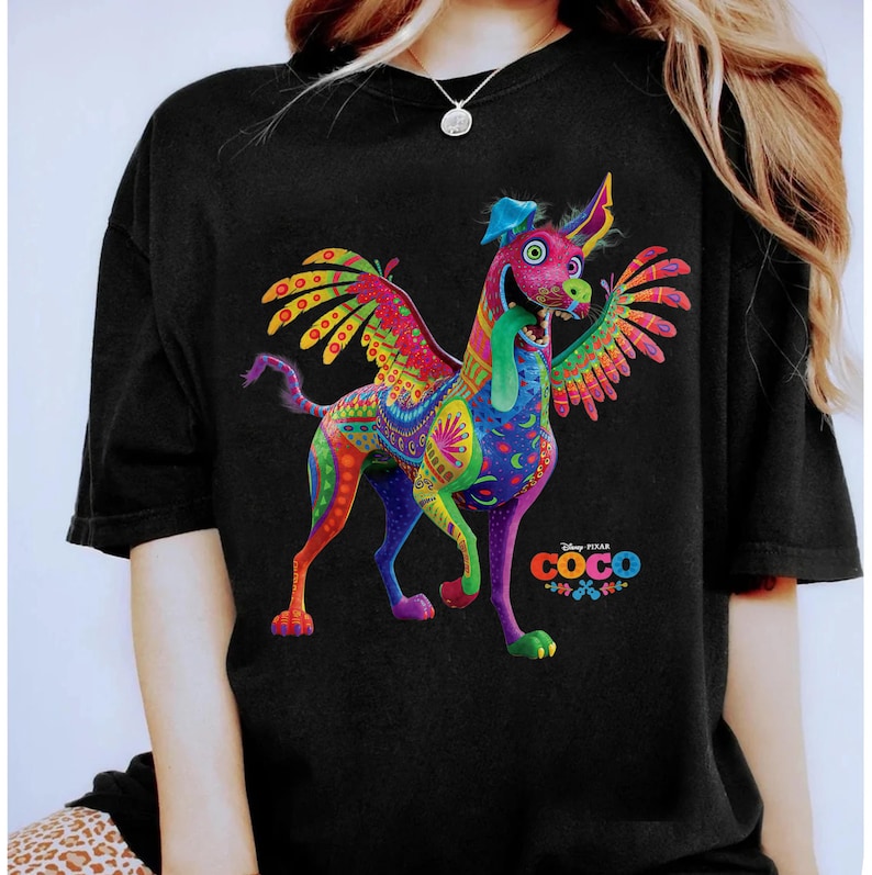 May include: A black short-sleeved t-shirt featuring a colorful graphic of Dante the alebrije dog from the movie Coco. The dog has wings, a long tongue, and intricate patterns in shades of pink, purple, yellow, and green. The Coco logo is visible.
