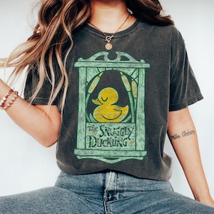 May include: A charcoal gray short-sleeve t-shirt featuring a graphic print of a yellow rubber duck inside a green wooden frame with cattails. The text "The Snuggly Duckling" is printed in dark green lettering at the bottom of the frame design.