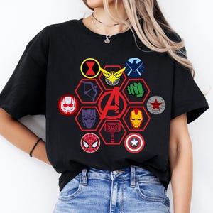 Puede incluir: Camiseta negra con un gráfico colorido del logotipo de los Vengadores y varios símbolos de personajes en un patrón de panal. El diseño incluye la máscara de Iron Man, el escudo del Capitán América y la máscara de Spider-Man.