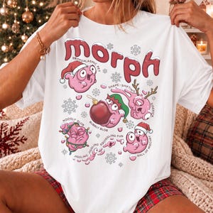 May include: A white t-shirt featuring a large red graphic with the text "MORPH" at the top. The design includes several pink, blob-like creatures in various holiday-themed poses, surrounded by small snowflakes and descriptive text labels.