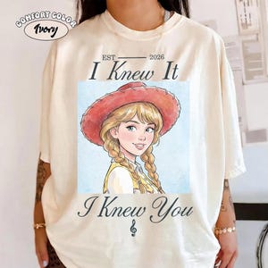 May include: An ivory t-shirt featuring a graphic of a cartoon character with blonde braided hair wearing a red cowboy hat. The text reads EST 2026 I Knew It I Knew You with a musical note symbol. A small oval label in the corner says COMFORT COLOR Ivory.