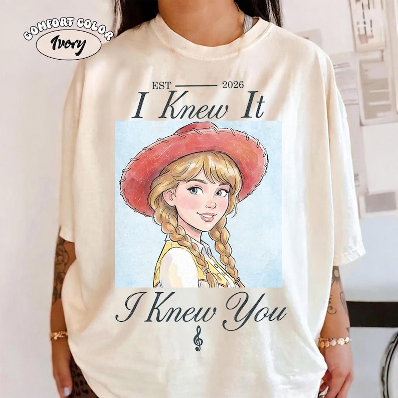 May include: An ivory t-shirt featuring a graphic of a cartoon character with blonde braided hair wearing a red cowboy hat. The text reads EST 2026 I Knew It I Knew You with a musical note symbol. A small oval label in the corner says COMFORT COLOR Ivory.