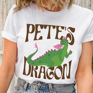 May include: White t-shirt with a brown graphic of a green dragon with pink accents and the text "Pete's Dragon".