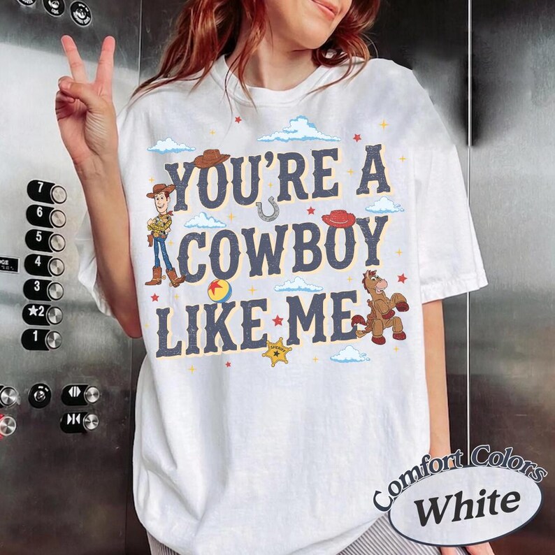 May include: A white t-shirt featuring the text "YOU'RE A COWBOY LIKE ME" in dark blue letters. The design includes illustrations of a cowboy character, a horse, a ball, a sheriff star, and small red stars, all surrounded by light blue clouds.