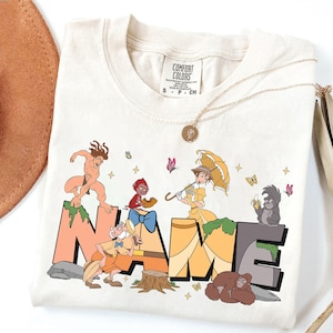 May include: Cream Comfort Colors t-shirt with a colourful graphic design. The design spells "NAME" in large, stylised letters with cartoon characters and butterflies. A gold necklace and a brown hat are also visible.