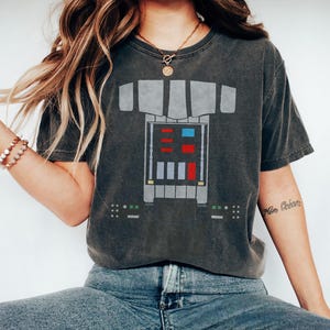 Op de afbeelding: Een antracietgrijs t-shirt met korte mouwen en een grafische print van een mechanische borstplaat. Het ontwerp bevat een centraal rechthoekig paneel met rode, blauwe en lichtgrijze vierkante accenten en kleinere bedieningspanelen.