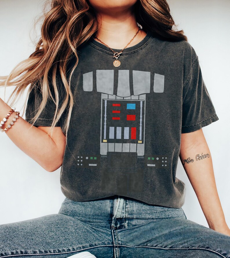Op de afbeelding: Een antracietgrijs t-shirt met korte mouwen en een grafische print van een mechanische borstplaat. Het ontwerp bevat een centraal rechthoekig paneel met rode, blauwe en lichtgrijze vierkante accenten en kleinere bedieningspanelen.