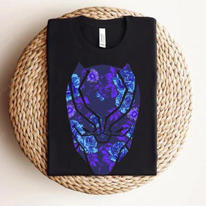Könnte beinhalten: Schwarzes T-Shirt mit einem floralen Panthermasken-Design in Blau- und Lilatönen. Die Grafik ist mittig platziert und kombiniert Superheldenästhetik mit floralen Elementen. Das T-Shirt ist auf einer gewebten Matte gefaltet.