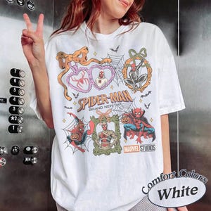 May include: A white t-shirt featuring a graphic print with the text Spider-Man Brand New Day and Marvel Studios. The design includes multiple illustrations of Spider-Man in various costumes, spiderwebs, bats, pumpkins, and decorative frames.