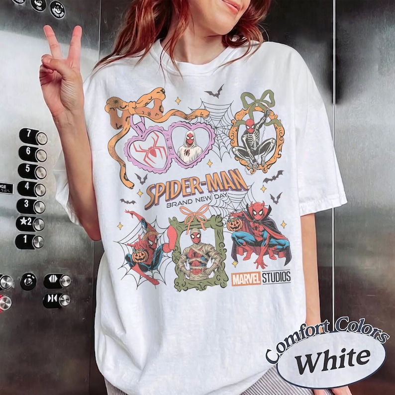 May include: A white t-shirt featuring a graphic print with the text Spider-Man Brand New Day and Marvel Studios. The design includes multiple illustrations of Spider-Man in various costumes, spiderwebs, bats, pumpkins, and decorative frames.