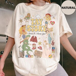 May include: A cream-coloured short-sleeve t-shirt featuring a graphic print with the text TAY STORY and I Knew It, I Knew You. The design includes multiple illustrated figures, a red cardigan, cowboy boots, a hat, and a sign reading Tay's Room Keep Out.