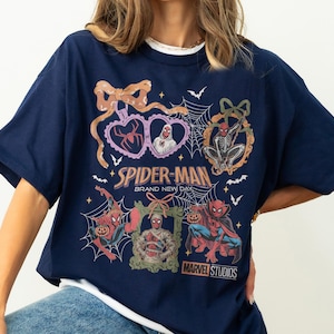 May include: A navy blue t-shirt featuring a graphic print of Spider-Man in various Halloween-themed costumes and poses. The design includes spider webs, bows, and frames, with the text Spider-Man Brand New Day and Marvel Studios printed on the front.