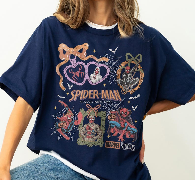 May include: A navy blue t-shirt featuring a graphic print of Spider-Man in various Halloween-themed costumes and poses. The design includes spider webs, bows, and frames, with the text Spider-Man Brand New Day and Marvel Studios printed on the front.