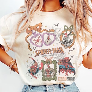 May include: A cream-colored t-shirt featuring a graphic print of Spider-Man in various Halloween-themed frames and poses. The design includes spiderwebs, bats, and the text Spider-Man Brand New Day and Marvel Studios in a colorful, vintage-inspired style.