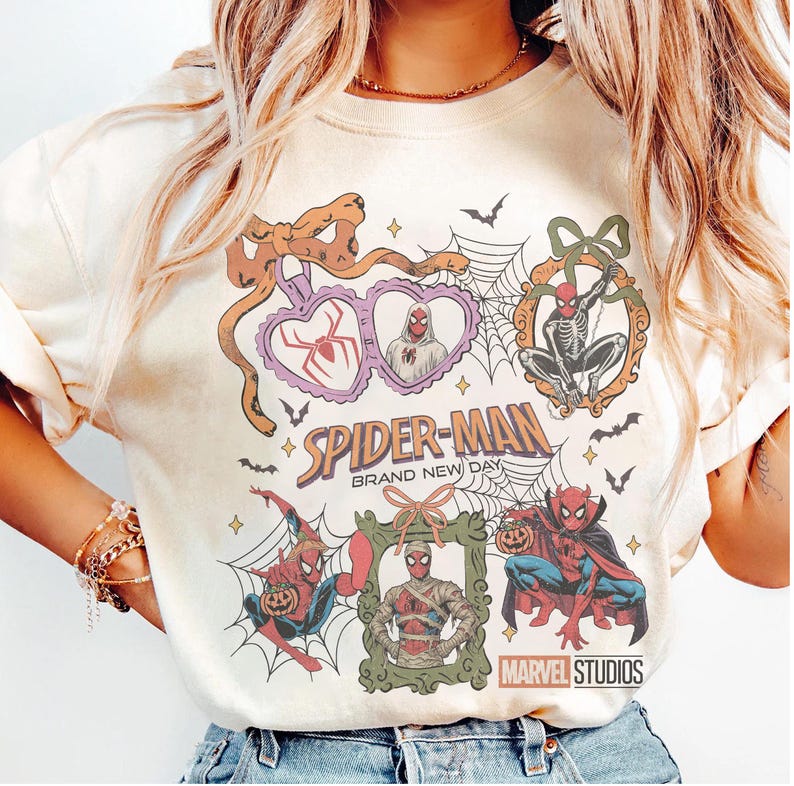 May include: A cream-colored t-shirt featuring a graphic print of Spider-Man in various Halloween-themed frames and poses. The design includes spiderwebs, bats, and the text Spider-Man Brand New Day and Marvel Studios in a colorful, vintage-inspired style.