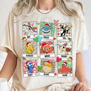 Puede incluir: Camiseta color crema con una cuadrícula de personajes de dibujos animados de la película "Buscando a Nemo" con gorros de Papá Noel. Cada personaje está en una imagen enmarcada con decoraciones navideñas y su nombre. Personajes: Ray, Bruce, Gill, Bubbles, Nemo, Squirt, Dory, Bloat y Hank.