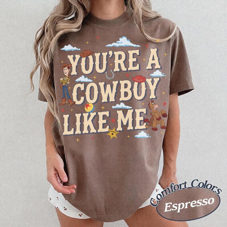 May include: A brown short-sleeve t-shirt featuring the text YOU'RE A COWBOY LIKE ME in cream lettering. The graphic includes a cartoon cowboy, a horse, a horseshoe, a star, a ball, and clouds. The shirt has a crew neckline and a relaxed fit.