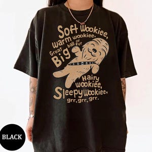 May include: Black t-shirt featuring a tan cartoon Wookiee graphic and text. The text reads "Soft Wookiee, warm wookiee, Great ball fur, Big, Hairy wookiee, Sleepy Wookiee, grr, grr, grr." A black circle with the word "BLACK" is in the lower left corner.