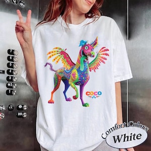 May include: A white t-shirt featuring a vibrant, multi-colored graphic of a winged alebrije dog with a long tongue. The design includes intricate patterns in shades of pink, blue, yellow, and green, with the word COCO printed in colorful letters.