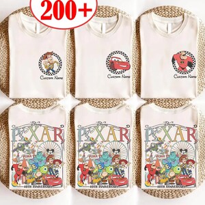 Puede incluir: Camisetas blancas con diseños de personajes de Disney Pixar. Los diseños incluyen a Woody, Rayo McQueen y Mr. Increíble en marcos circulares, y un gráfico más grande del 40 aniversario de Pixar. El texto "Custom Name" aparece debajo de algunos diseños.