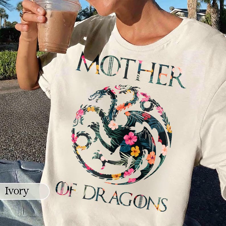 May include: An ivory t-shirt featuring the text MOTHER OF DRAGONS in a dark green font. Centered below is a circular graphic of a three-headed dragon filled with a colourful tropical floral pattern of pink, yellow, and green leaves.