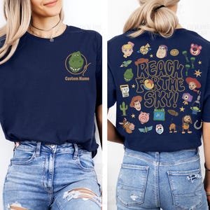 May include: A navy blue t-shirt featuring a small graphic of a green dinosaur head inside a lasso on the front left chest with the text Custom Name below it, and a large back print with various cartoon characters and the text REACH FOR THE SKY!