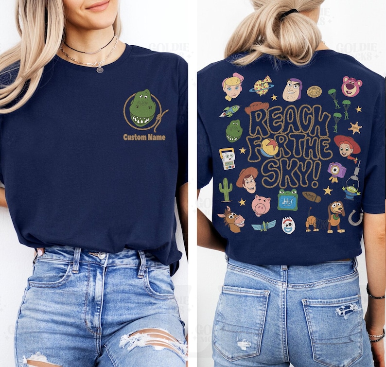 May include: A navy blue t-shirt featuring a small graphic of a green dinosaur head inside a lasso on the front left chest with the text Custom Name below it, and a large back print with various cartoon characters and the text REACH FOR THE SKY!