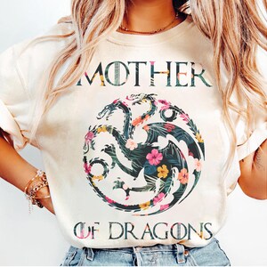 May include: A cream-coloured t-shirt featuring the text MOTHER OF DRAGONS in a dark font. Centered is a circular graphic of a three-headed dragon filled with a tropical pattern of green palm leaves and vibrant pink, yellow, and orange hibiscus flowers.